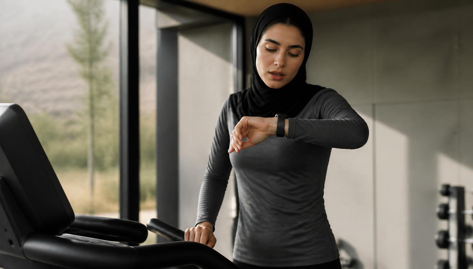 Une femme d'origine moyen-orientale en tenue de sport sobre consulte sa montre connectée après une séance de course, dans une salle d'entraînement moderne éclairée par la lumière naturelle
